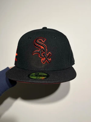 Svart och röd Chicago New Era 59FIFTY keps - Säljer en svart New Era 59FIFTY keps med broderad röd och svart White Sox-logga framtill och frihetsgudinnan på sidan. Kepsen har platt skärm och klassisk passform, med röda detaljer längs kanten. Perfekt för dig som gillar streetwear och sportiga vibes.