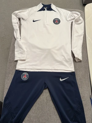 PSG x Nike träningsset marinblå/vit - Säljer ett fett PSG träningsset från Nike med långärmad vit tröja och marinblå byxor. Båda delarna har Paris Saint-Germain-logga och Nike swoosh. Tröjan har half zip och byxorna är smala i modellen. Perfekt för dig som älskar fotboll och vill ha stil på träningen.