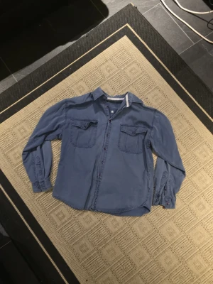 Blå skjorta med fickor och knappar - Säljer en blå skjorta i bomull med två stora bröstfickor och röda knappar. Skjortan har klassisk krage och lång ärm med knapp vid manschetten. Passformen är normal och materialet känns mjukt och skönt mot huden. Perfekt att styla med jeans eller chinos.