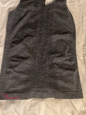 Grå träningstopp från Panos - Säljer en gråmelerad linnetopp från Panos med snyggt mönstrad mesh-panel längs mitten. Toppen har racerback och ärmlös design, perfekt för gymmet eller löpning. Materialet är mjukt och stretchigt syntet för maximal komfort. Diskret rosa logga nertill.
