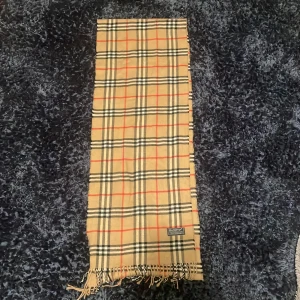 Rutig beige halsduk från Burberrys - Säljer en klassisk beige halsduk från Burberrys med det ikoniska rutiga mönstret i svart, vitt och rött. Halsduken är tillverkad i 100% lammull och har fransar i ändarna. Perfekt accessoar för att lyfta din outfit med lite brittisk stil.