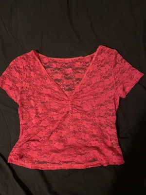 Rosa  spets topp med v-ringning - Säljer en rosa , kortärmad topp i genombruten spets med v-ringning och rynkad detalj framtill. Toppen är croppad och har ett blommigt spetsmönster som ger en romantisk vibe. Perfekt att styla med höga jeans eller kjol.
