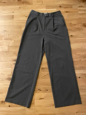 Grå kostymbyxor från Djerf Avenue (M)  - Snygga grå kostymbyxor från Djerf Avenue med hög midja och breda ben. Byxorna har klassiska pressveck, bälteshällor och fickor fram samt en bakficka. Materialet är mjukt och har en elegant känsla, perfekt för en clean och stilren look. (Sista bilderna är lånade från Djerf Avenues hemsida) 