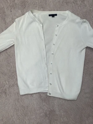 Vit kofta från Tommy Hilfiger - Snygg vit kofta från Tommy Hilfiger med rund hals och silverfärgade knappar där det står Tommy Hilfiger. Långärmad modell med diskret logga på ärmen. Mjuk och tunn stickad kvalitet som passar perfekt till lager-på-lager. Ribbad kant vid ärmslut och nederkant.