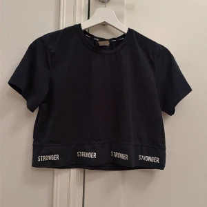 Svart croppad topp från Stronger - Säljer en svart croppad topp från Stronger med korta ärmar och rund hals. Nederkanten har vit text med märket 'STRONGER' tryckt flera gånger. Perfekt för träning eller chill dagar.