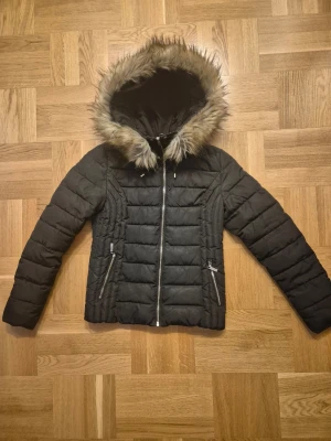 Svart pufferjacka från Roco Barocco stl 38 - Svart pufferjacka med avtagbar huva i fuskpäls från Roco Barocco. Jackan har quiltad design, dragkedjor på fickorna och leopardmönstrat foder. Perfekt för kalla dagar med sin fluffiga känsla och snygga detaljer. Mycket bra skick!