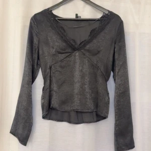 Grå långärmad omlottopp med spets - Snygg grå långärmad topp i glansigt material med v-ringning och spetsdetaljer längs halsen. Omlott-design framtill ger en schysst passform och lite extra stil. Perfekt att matcha med jeans eller kjol för en trendig look.