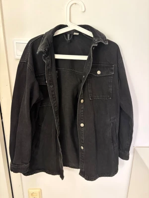 Svart jeansjacka från H&M Divided XS - Säljer en svart jeansjacka från H&M Divided i storlek XS. Jackan har klassisk krage, knappstängning framtill, bröstficka med lock och knapp samt sidofickor. Perfekt oversized look och passar till många olika stilar.