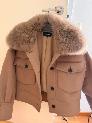 Beige pälsjacka från Meotine XS/S - Säljer en beige pälsjacka från Meotine med stor fluffig krage i pälsimitation. Jackan har två stora fickor med lock och svarta knappar framtill. Modellen är croppad och har en loose passform, perfekt för att ge en cool vibe till din outfit.
