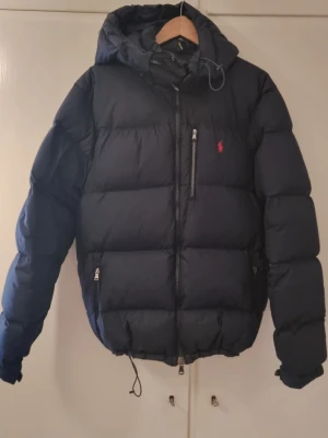 Mörkblå dunjacka från Polo Ralph Lauren - Large - Mörkblå dunjacka från Polo Ralph Lauren – The Gorham Down Jacket (uppfattar jag). Jacka med huva, dragkedja och röd Polo-logga på bröstet. Flera praktiska fickor med dragkedja (3 ytterfickor + 1 innerficka) samt justerbar nederkant med snörning. Ett pyttelitet hål (se bild 2), enkelt att laga – i övrigt mycket fint skick. Säljs p.g.a. felköp, för stor för sonen. Storlekslapp saknas, mått motsvarar Large (rymlig modell): bröstvidd 130 cm, axel 53 cm, ärm 96,5 cm, nederkant 125 cm (reglerbar).
