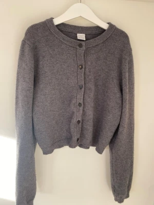 Grå kofta från Lindex med knappar. - Mysig grå kofta från Lindex i storlek 146/152. Koftan har rund hals, långa ärmar och svarta knappar framtill. Materialet är stickat och känns mjukt mot huden. Perfekt för lager-på-lager under kyliga dagar. Använd få tal gånger. Som ny.