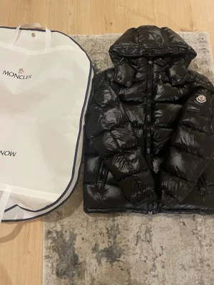 Moncler jacka - Säljer en svart pufferjacka från Moncler med glansig finish, huva och dragkedja framtill. Jackan har Moncler-logga på ärmen och praktiska fickor med dragkedja. Perfekt för kalla dagar och har en riktigt snygg streetstil.