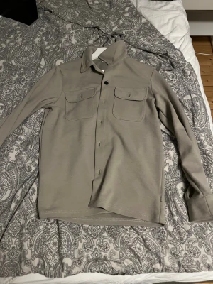 Beige overshirt från Only & Sons - Snygg beige overshirt från Only & Sons med klassisk krage och två stora bröstfickor med lock. Jackan har knappar framtill och är långärmad, perfekt att slänga på över en t-shirt för en chill look.