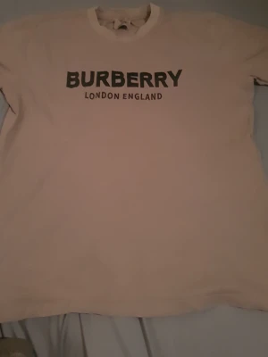 Beige Burberry t-shirt med tryck - Säljer en beige t-shirt från Burberry i storlek XXL med svart Burberry London England-tryck på bröstet. T-shirten har rund hals och är kortärmad, tillverkad i 100% bomull för skön och mjuk känsla. Perfekt för dig som gillar klassiska designer och streetstyle.