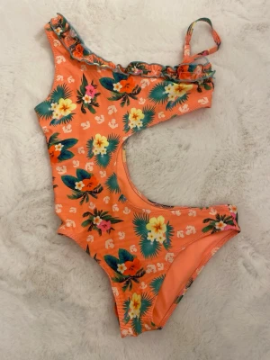 Orange blommig baddräkt med volang - Säljer en snygg orange baddräkt med tropiskt blommigt mönster och volangdetalj längs axeln. Baddräkten har en cool cut-out design och en axelrem. Perfekt för strandhäng och poolparty. har använt 1 gång!!!!!!!!!!!!!!!!!!
