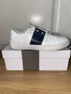 Valentino Garavani vita sneakers - Säljer ett par vita Valentino Garavani sneakers med bred mörkblå rand på sidan och coola pyramidnitar runt hälen. Skorna har snörning, rund tå och platt sula. Tillverkade i skinn med vita skosnören och detaljer i blått. Perfekt för dig som gillar stilrena designer.