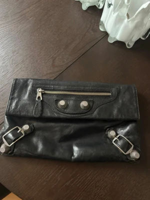 Svart kuvertväska från Balenciaga - Säljer en svart kuvertväska från Balenciaga i mjukt skinn med coola silverfärgade detaljer och spännen framtill. Väskan har en dragkedja på framsidan och magnetknäppning. Perfekt storlek för det viktigaste och har en stilren, edgy look. Köpt secondhand så vet ej äkthet men har serienummer i sig! Den ena niten håller på att gå av men går säkert att fixa men inget som syns eller försämrar användningen!