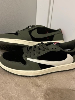 Nike Air Jordan 1 Low Travis Scott - Nike Air Jordan 1 Low Travis Scott i olivgrönt och svart med vit swoosh. Skorna har röda broderade detaljer på hälen och klassisk snörning. Materialet är en mix av skinn och mocka, med platt sula och rund tå. Perfekt för dig som gillar streetwear och sneakers med unik design.