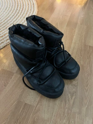 Svarta vinterskor med platt sula och vaddering  - Svarta vinterskor med tjock platt sula och snörning framtill. insidan är fodrad med mjukt pälsliknande tyg för extra värme. Skorna har en rund tå och en chunky siluett som sticker ut.