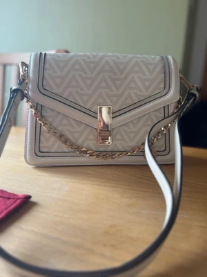 Vit axelväska från Aldo med kedja - Snygg vit axelväska från Aldo med geometriskt mönster, guldfärgad kedjedetalj och lås framtill. Väskan har justerbar axelrem och flera fack inuti för smart organisering. Perfekt storlek för det viktigaste och stilren look med lyxiga detaljer.