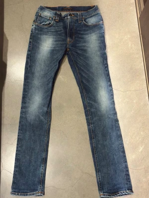 Blå jeans från Nudie Jeans  - Säljer ett par klassiska blå jeans från Nudie Jeans Co med snygga slitningar och kontrastsömmar. Modellen har raka ben, fem fickor och orange broderi på bakfickorna. Jeansen är tillverkade i bomull och har en normal passform med medelhög midja. Liknande fades som på selvagde jeans💥  W:29 L:32