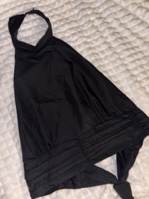 Svart omlott halterneck topp - Säljer en svart halterneck topp med omlott och knyt i ryggen. Toppen är ärmlös och har en bred kant nedtill som ger en snygg siluett. Materialet känns som bomull eller polyester och toppen har öppen rygg. 