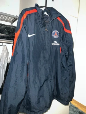 PSG vindjacka Nike XL - Säljer en mörkblå PSG vindjacka från Nike i storlek XL. Jackan har röda detaljer längs axlarna, dragkedja framtill och PSG-logga samt Fly Emirates-tryck på bröstet. Perfekt för dig som gillar fotboll och vill ha en sportig look.