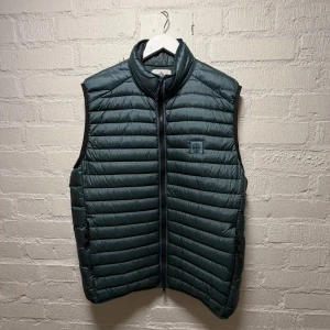 Stone Island Dunväst - Säljer en mörkgrön dunväst från Stone Island med quiltad design och dragkedja framtill. Västen har ståkrage och ett broderat Stone Island-märke på bröstet. Perfekt för lager-på-lager och snygg till streetwear. Färgtonen är mörkgrön och materialet är dun.