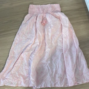 Ljusrosa långkjol med tofsdetalj - Säljer en ljusrosa långkjol med paisleyinspirerat mönster. Kjolen har bred smock i midjan och dekorativa tofsar med pärlor framtill. Materialet känns lätt och luftigt, perfekt för varma dagar. Passar dig som gillar boho-vibe och mjuka färger.