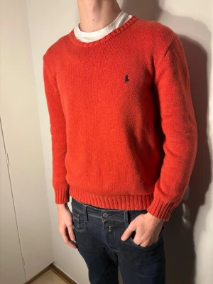 Röd stickad tröja från Ralph Lauren - Säljer en snygg röd stickad tröja från Ralph Lauren med klassisk rund hals och diskret logga broderad på bröstet. Tröjan har ribbade muddar vid ärmslut och nederkant samt långärmad passform. Perfekt för dig som gillar stilrena och tidlösa plagg.