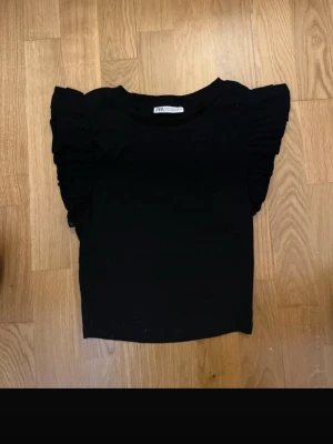 Svart topp med volangärmar från Zara - Säljer en svart topp från Zara i storlek M. Toppen har korta ärmar med snygga volangdetaljer som ger en cool och trendig look. Materialet är mjukt och ribbat, vilket gör den skön att bära. Perfekt att styla med jeans eller kjol för en enkel men chic outfit.