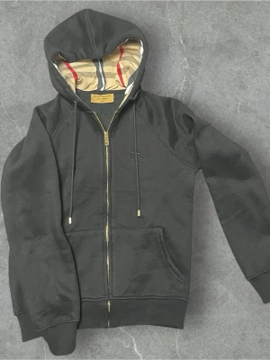 Burberry Zip-Svart - Svart zip hoodie från Burberry i klassisk och stilren design. Hoodie med hel dragkedja framtill, mjukt och följsamt material samt huva med justerbara snören. Diskreta Burberry-detaljer som ger ett exklusivt intryck utan att bli för mycket. Plagget är väl omhändertaget och i fint skick – inga hål, fläckar eller större slitage. Bilderna visar både helhet, material, dragkedja och detaljer för en tydlig bild av skick och kvalitet. Liten i storlek, passar small.  