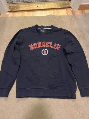 Mörkblå sweatshirt från Bondelid S - Klassisk mörkblå sweatshirt från Bondelid med röd logga och vit detalj framtill. Tröjan har rund halsringning, ribbade muddar och är tillverkad i mjuk bomull. Perfekt för en chill och stilren look.