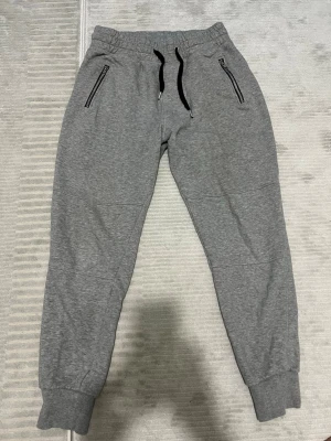 Grå mjukisbyxor från H&M Divided M - Grå mjukisbyxor med svarta snören i midjan och dragkedjor på fickorna fram. Byxorna har muddar vid bensluten och två bakfickor. Tillverkade i en mjuk blandning av bomull, polyester och viskos. Perfekta för chill och häng.