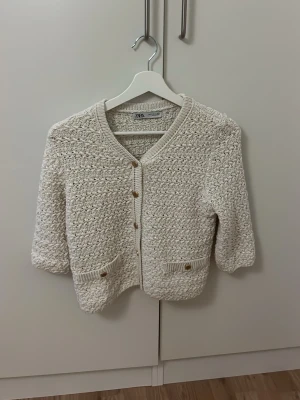 Beige stickad kofta från Zara - Säljer en beige stickad kofta från Zara med glittriga inslag i materialet. Koftan har v-ringad hals, trekvartsärmar och snygga knappar framtill samt två små fickor med knappdetalj. Perfekt för dig som gillar en chill och trendig look.
