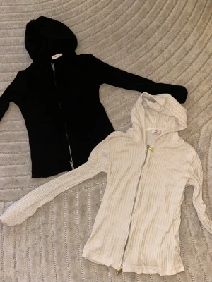 Två tunna huvtröjor från Nelly med ribbat material. - Säljs tillsammans. En beige och en svart tunn huvtröja från Nelly, båda i storlek S. Nytt skick. Dragkedjan kan justeras både uppifrån och nedifrån. 