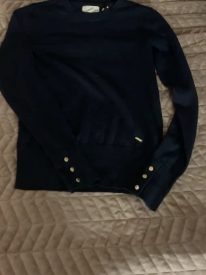 Mörkblå stickad tröja Holly & Whyte - Snygg mörkblå stickad tröja från Holly & Whyte by Lindex i storlek XS. Tröjan har långa ärmar och coola guldfärgade knappar vid ärmsluten som ger en extra touch. Perfekt till jeans eller kjol för en clean look.