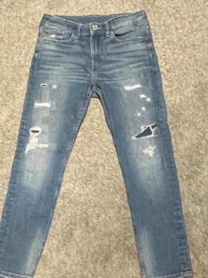 Blå skinny jeans med slitningar - Säljer ett par blå skinny jeans med snygga slitningar och hål på framsidan. Jeansen har klassisk femficksdesign, knapp och dragkedja i midjan. Materialet är jeans och passformen är tight med smal siluett. Perfekt för dig som gillar en trendig och avslappnad stil. Eftersom dom inte passar mig längre 