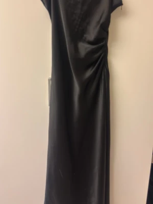 Svart långklänning i satin-look från Zara  - Säljer en svart långklänning med glansig satin-look och snygg rynkning i sidan. Klänningen har korta ärmar och en rak, elegant siluett. Perfekt för dig som vill ha en stilren och trendig outfit.