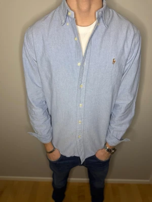 Ljusblå skjorta från Polo Ralph Lauren - Snygg ljusblå skjorta från Polo Ralph Lauren med klassisk button-down krage och broderad logga på bröstet. Custom fit-modell med långa ärmar och vita knappar framtill. Tillverkad i mjuk bomull som känns skön mot huden. Storlek L. Skick 8/10. Vi kan gå ner i pris vid snabb och smidig affär. // Studio II Resell 