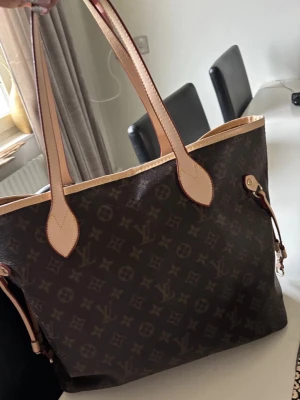 Brun väska  - Säljer en klassisk brun väska  med ikoniskt monogrammönster. Väskan har långa handtag i ljusbeige läder och detaljer i samma färg. Perfekt storlek för att få plats med allt du behöver och riktigt snygg att bära på axeln.