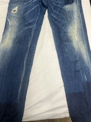 Dsquared 2 Blåa Jeans - Säljer ett par blå jeans med coola slitningar och blekta detaljer på både fram- och baksida. Som ni ser så är jeansen gjorda av DsQuared2. Jeansen har raka ben och klassiska fickor bak. Materialet är jeans och färgen är mörkblå med ljusare partier. Perfekt för dig som gillar en avslappnad streetstil.