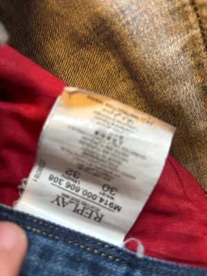 Replay Anbass jeans blå, storlek 30/32 - Säljer ett par Replay Anbass jeans i klassisk blå tvätt med snygga slitningar och kontrastsömmar. Modellen har normal passform och raka ben, med röda Replay-detaljer vid fickan och insidan. Jeansen är tillverkade i bomull och har en stilren look.