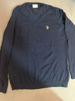 Marinblå stickad tröja U.S. Polo Assn. - Säljer en marinblå stickad tröja från U.S. Polo Assn. med v-ringad hals och broderad logga på bröstet. Tröjan har långa ärmar och ribbade muddar vid ärmslut och nederkant. Perfekt för dig som gillar klassisk och sportig stil.jääte fin och stilig du kan returnera direkt om du inte gillar 