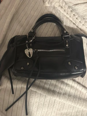 Svart vintage väska - Snygg svart handväska i skinn med coola silverdetaljer och dragkedja framtill. Väskan har dubbla handtag, dekorativa nitar och ett hängande hjärtberlock med vingar. Långa remmar och extra fack på utsidan ger en edgy vibe, knappt använt. 