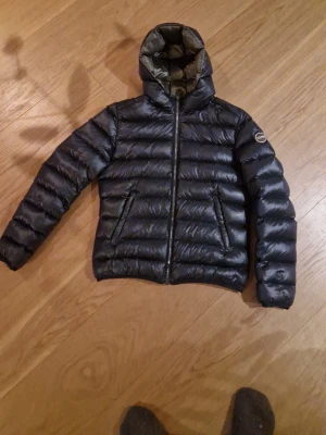 Mörkblå dunjacka från Colmar - Säljer en snygg mörkblå dunjacka från Colmar med huva och dragkedja framtill. Jackan har ett quiltat mönster och två fickor med dragkedja. Perfekt för kalla dagar och har en cool, sportig vibe. Passar dig som gillar stil och komfort.