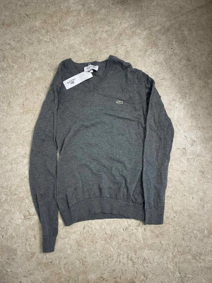 Grå v-ringad tröja från Lacoste - Snygg grå v-ringad tröja från Lacoste med den klassiska krokodilloggan broderad på bröstet. Tröjan har lång ärm, ribbade muddar och är tillverkad i mjuk bomull. Perfekt för dig som gillar stilrena och enkla plagg med en touch av sportig vibe.