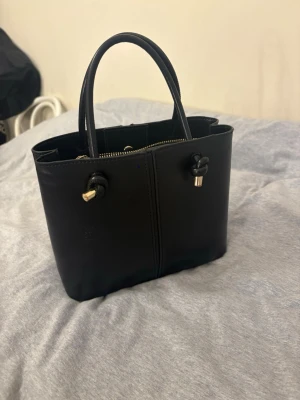 Svart handväska med gulddetaljer - Snygg svart handväska i skinn med stilrena guldfärgade detaljer och praktiska handtag. 