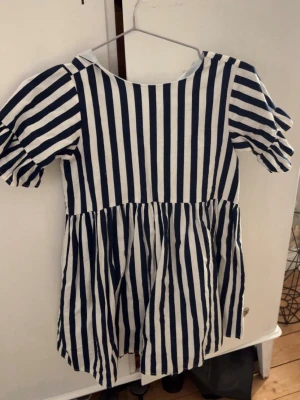 Randig kortklänning från POMPdeLUX - Säljer en söt kortklänning från POMPdeLUX i storlek 122/128. Klänningen har marinblå och vita vertikala ränder, puffiga kortärmar och en rund halsringning. Tillverkad i mjuk bomull som är skön mot huden. Perfekt för dig som gillar klassisk och lekfull stil.