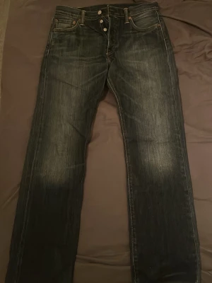 Levi's 501 mörkblå jeans W30 L30 - Klassiska Levi's 501 jeans i mörkblå tvätt med raka ben och knappgylf. Jeansen har fem fickor, kontrastsömmar och den ikoniska röda Levi's-lappen bak. Perfekta för dig som gillar tidlös stil och vill ha ett par slitstarka jeans i bomull.